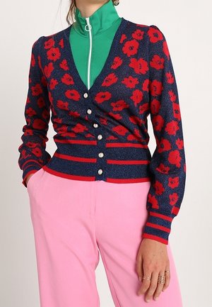 Vrouw in een marineblauwe cardigan met rode bloemenpatronen over een groene zip-up top, gecombineerd met hoogwaisted roze broek.