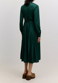 Langärmliges grünes Kleid mit einem plissierten Rock, tailliertem Schnitt und Knopfmanschetten. Kombiniert mit braunen, klobigen Clogs.