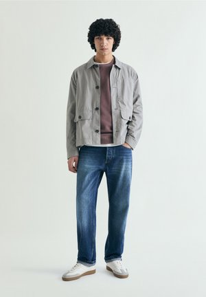 Young man with curly hair wears gray button jacket, brown shirt, blue jeans, and white sneakers, standing against plain light background.