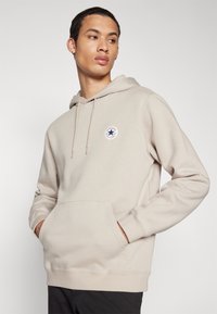 Beige hettegenser laget av bomullsblanding, har en frontlomme, snorhette og en sirkulær logopatch på venstre brystområde.