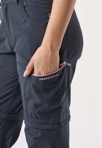 Granatowe spodnie outdoorowe z kieszenią na zamek, różowym akcentem zamka i ozdobnym przeszyciem. Materiał wygląda na gładki i odporny na warunki atmosferyczne.