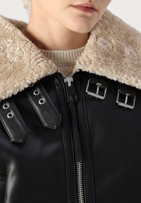 Veste en cuir noir avec un col en fausse fourrure beige, fermeture éclair, accessoires en métal argenté, et éléments décoratifs avec œillets et boucles.
