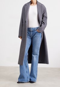 Abrigo largo de lana gris con solapa y bolsillos frontales, combinado con jeans acampanados en azul claro y una blusa blanca ajustada.