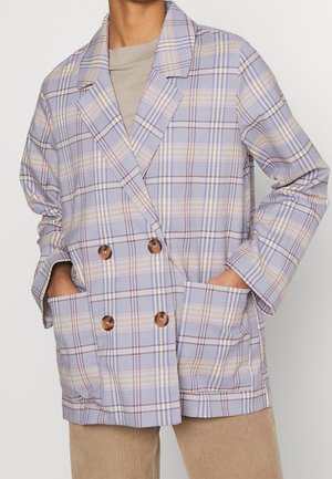 Personne portant un blazer croisé à carreaux violet clair et beige avec de gros boutons marron et les mains dans les poches avant.