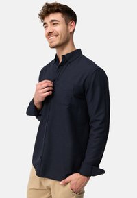 Camisa de botones azul marino hecha de una tela de textura suave. Presenta un bolsillo en el pecho y un cuello abotonado. Mangas largas enrolladas en los puños.