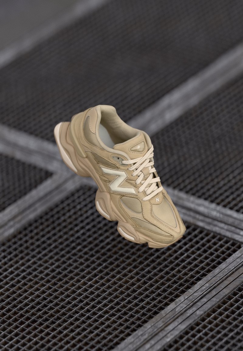 Baskets beige New Balance avec une semelle épaisse et le logo blanc "N" posées sur une surface en grille métallique avec des lignes diagonales.