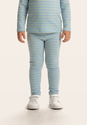 Lapsi, jolla vaaleansininen ja keltainen raidallinen pitkähihaisen yläosa ja yhteen sopivat leggingsit valkoisilla tennareilla, seisoo vaalealla taustalla.