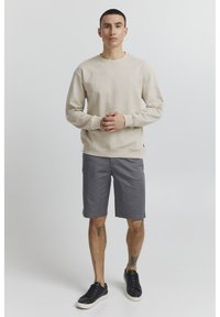 Sudadera beige clara de manga larga, combinada con shorts grises y zapatillas negras. El modelo está de pie, con las manos entrelazadas, sobre un fondo sencillo.