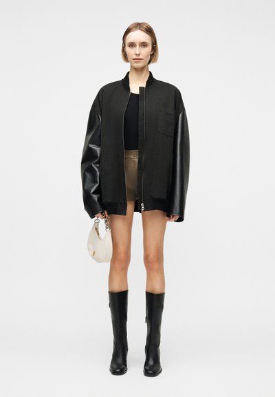 Νεαρή γυναίκα με υπερμεγέθη μαύρο μπουφάν με δερμάτινα μανίκια, μαύρο τοπ, χακί σορτς, μαύρες μπότες μέχρι το γόνατο, κρατώντας λευκή τσάντα.