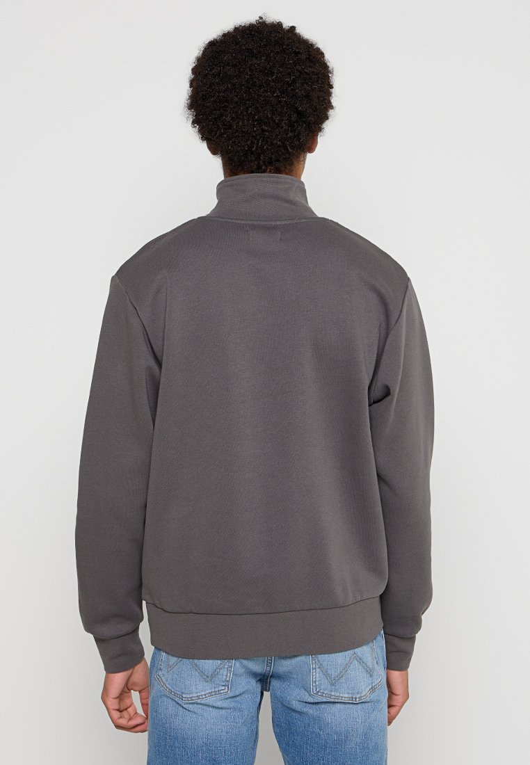 Sudadera gris con un cuello alto, puños y dobladillo acanalados. Textura suave, corte informal y diseño minimalista sin logos visibles.