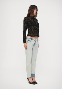 Woman wearing a black long-sleeve floral lace blouse, light blue faded straight jeans, and black pointed high heels, standing sideways.
