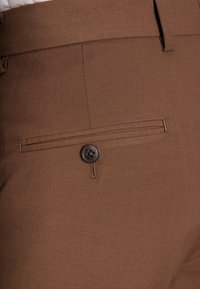 Brązowe spodnie z gładkiej tkaniny; posiadają zapięcie na guzik, detale szwów oraz pojedynczą tylną kieszeń z poziomym wejściem.