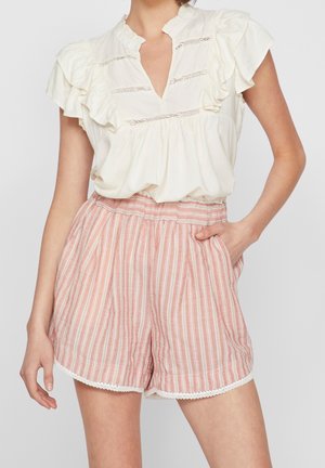 Femme portant un chemisier crème à volants rentré dans un short taille haute rayé rose et blanc avec bordure en dentelle, une main dans la poche.