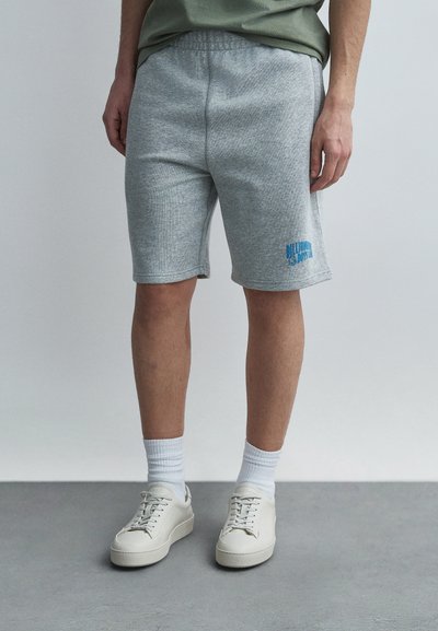 Shorts gris en coton avec une ceinture élastique, ornés d'un logo graphique bleu. Associés à des baskets montantes blanches et des chaussettes côtelées.