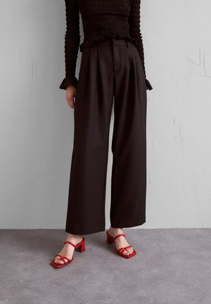 Kvinde iført sorte vidde bukser med høj talje, sort langærmet top og røde sandaler med blokhæl og stropper på grå gulv med hvid væg.