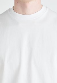 Tricou din bumbac alb simplu, cu mânecă scurtă și guler rotund; textură netedă și detalii minime, potrivit pentru îmbrăcăminte casuală.