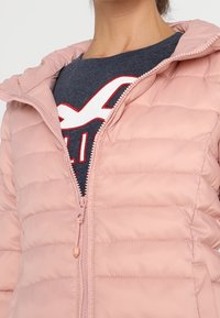 Doudoune rose clair avec texture matelassée horizontale, dotée d'une fermeture éclair frontale et d'une capuche. En dessous, une chemise bleu foncé avec des motifs rouges.