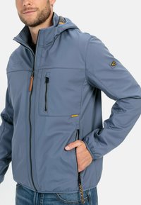Lys grå softshell-jakke med hætte, lynlåslukning foran og brystlomme. Har orange detaljer og en glat overflade.