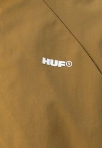 Brun vindjakke med en glat tekstur, der har et hvidt "HUF" logo på venstre side. Minimalistisk design, uden yderligere mønstre eller accenter.