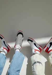 Deux paires de jambes portant des baskets blanches, noires et rouges avec des lacets, l'une dans un jean bleu clair et l'autre dans un pantalon tie-dye blanc, sur un fond uni.