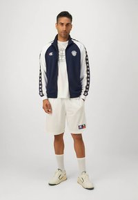 Homem a usar uma jaqueta com fecho éclair azul-marinho e branca, camiseta branca com um gráfico de basquetebol, calções brancos, meias e ténis, de pé contra um fundo simples.