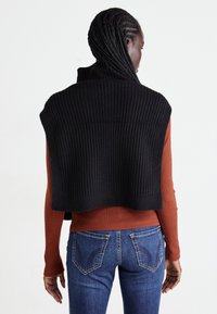 Frau mit geflochtenem Haar, die einen schwarzen gerippten ärmellosen Pullover über einem rostfarbenen langärmeligen Top und blauen Jeans trägt, von hinten gesehen.