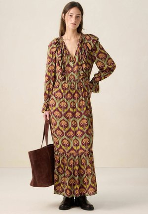 Robe longue bordeaux à motif floral jaune et vert, avec des épaules à volants et une taille froncée. Le modèle tient un sac fourre-tout marron.