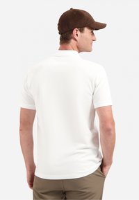 Witte polo met korte mouwen en een relaxed fit, met een klassieke kraag. Gecombineerd met een bruine pet, gezien vanuit de achterkant.