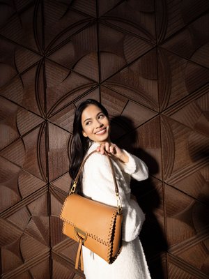 Brown leather handbag features a stitched edge, a gold-tone clasp, and a detachable shoulder strap. Textured background enhances visibility.