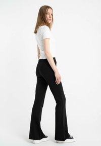 Czarne spodnie dzwony z elastycznego materiału, zestawione z dopasowaną białą koszulką. Modelka stoi bokiem na jednolitym tle.