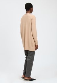 Beige gebreide cardigan met lange mouwen en zijopeningen, gecombineerd met donkergrijze getailleerde broek en zwarte instappers.