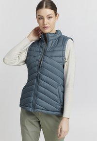Gilet en duvet bleu avec un col montant, design matelassé, poches latérales et fermeture éclair à l'avant. Le modèle porte un haut à manches longues de couleur crème.