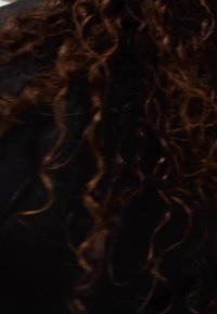 Kadeřavé vlasy střední délky s tmavohnědými odstíny a světlejšími zvýrazněními, vykazující texturovaný, objemný vzhled na černém pozadí.