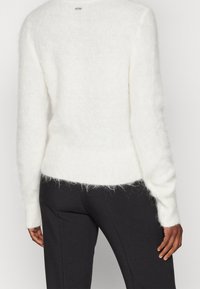 Skickad vit fluffig tröja med ribbad nederkant och ärmslut, kombinerad med svarta byxor, visad bakifrån på en stående person.