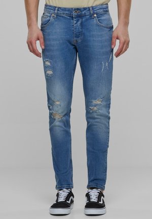Jean en denim bleu coupe skinny avec zones déchirées. Comprend des poches, une fermeture à bouton et une légère décoloration. Accentué par des baskets noires.