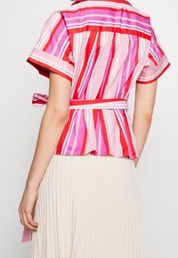 Chemise à manches courtes avec des rayures verticales multicolores en rouge, rose et violet. Comprend une taille à nouer et un tissu texturé.