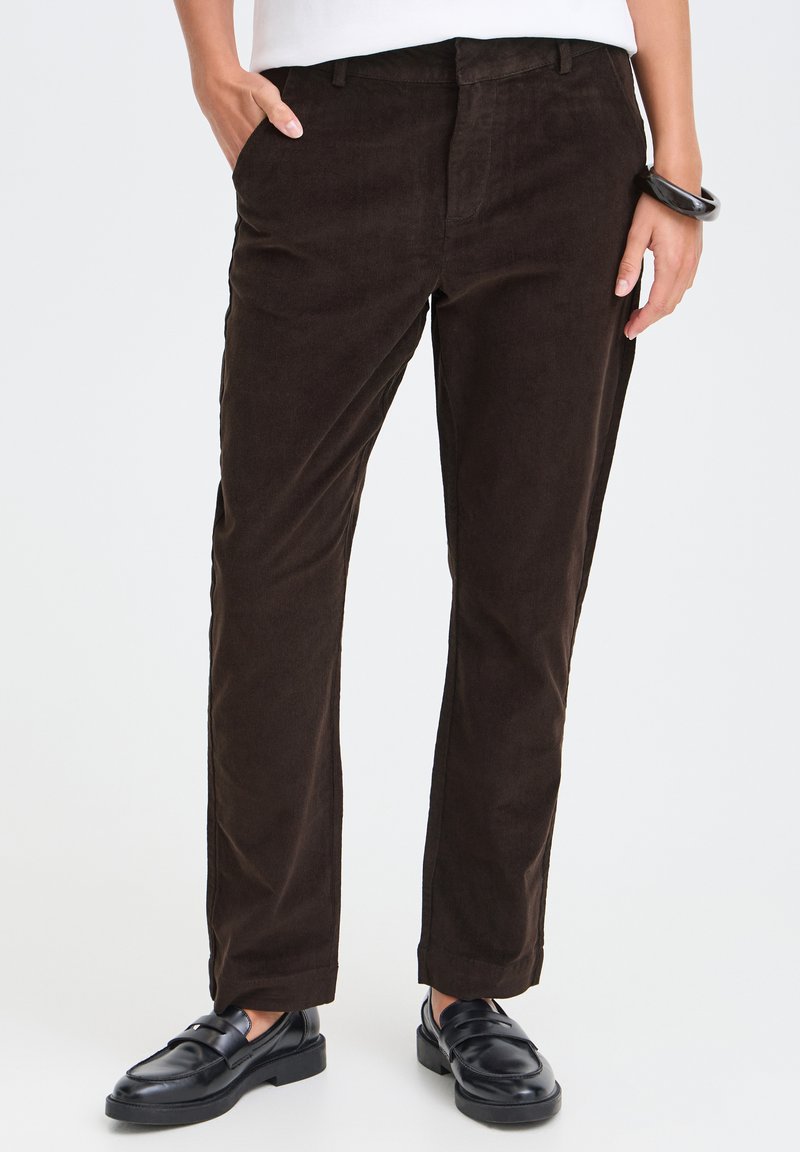 Pantalon droit marron foncé porté avec des mocassins noirs, une main dans la poche, l'autre avec un bracelet noir.