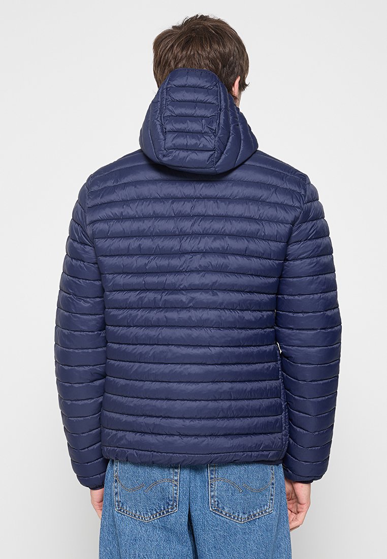 Mężczyzna w granatowej, pikowanej kurtce z kapturem i niebieskich dżinsach, pokazany od tyłu na jednolitym tle.