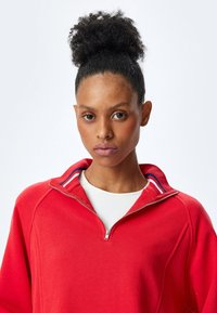 Czerwony sweter z kołnierzem na zamek i białą wewnętrzną warstwą. Posiada ribbrowany kołnierz w niebieskie i czerwone paski. Gładka tekstura materiału.