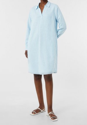 Mujer que lleva un vestido de rayas azul claro, de manga larga y largo hasta la rodilla, con sandalias blancas abiertas en la puntera, de pie sobre un fondo blanco.