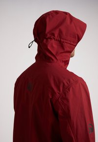Rode waterdichte jas met een aansluitende capuchon, elastische trekkoord en logodetail op de schouder. Lichtgewicht stof met een gladde textuur.