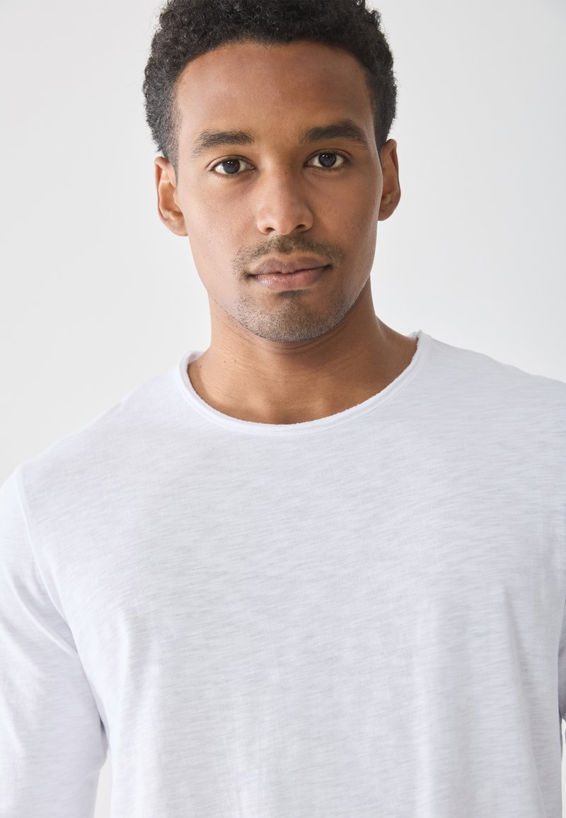 Biała koszulka z długim rękawem i okrągłym dekoltem, wykonana z miękkiego, teksturowanego materiału o minimalistycznym wzorze. Zbliżenie na górną część ciała.