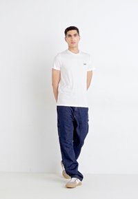 T-shirt en coton blanc avec un petit logo noir sur la poitrine, associé à un pantalon cargo bleu marine et des baskets beige clair. Design simple.