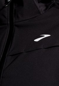 Juodas švarkas, pagamintas iš lygaus, lengvo audinio, su aukštu apykakle, priekyje užtrauktuku ir baltu logotipo akcentu. Dėl švaraus, minimalistinio dizaino.