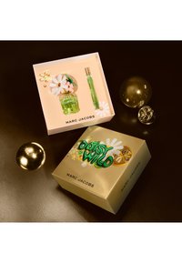 Groene glazen parfumfles met een bloemenkap, vergezeld van een reisspray. De omringende verpakking is roze en goud, met bloemaccenten.