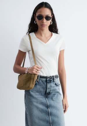 Mujer con gafas de sol ovaladas negras, camiseta blanca con cuello en V, falda de mezclilla, llevando un bolso marrón de hombro, de pie frente a un fondo liso.