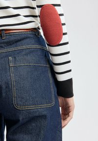 Gestreepte trui met een rode elleboogpatch, gecombineerd met blauwe spijkerbroek met opvallende achterzakken en gele stiksels.