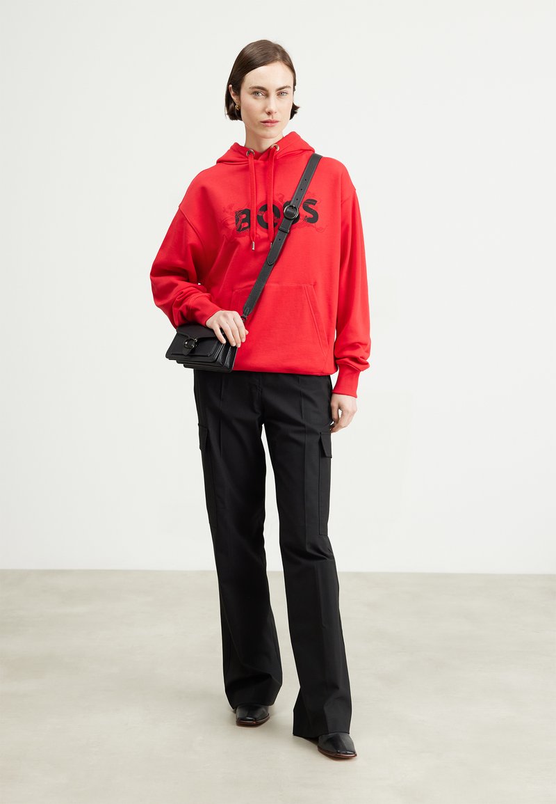 Sweatshirt rouge avec "BOSS" imprimé en noir, associé à un pantalon cargo noir. Le modèle porte un petit sac bandoulière noir.