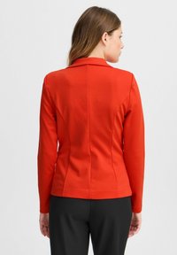 Veste rouge ajustée avec un col cranté, des manches longues et une texture lisse. Les coutures contournées le long du dos renforcent son aspect soigné.