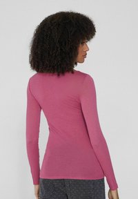 Žena s kudrnatými vlasy, oblečená v přiléhavém růžovém topu s dlouhými rukávy a tmavých vzorovaných džínách, otočená mírně na bok před jednolitým pozadím.